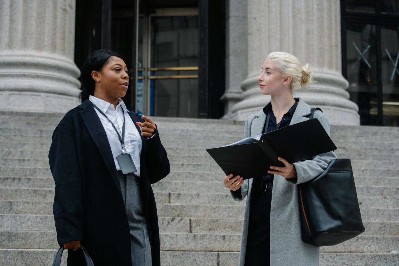 Two professional women in discussion on outdoor courthouse steps, holding documents.